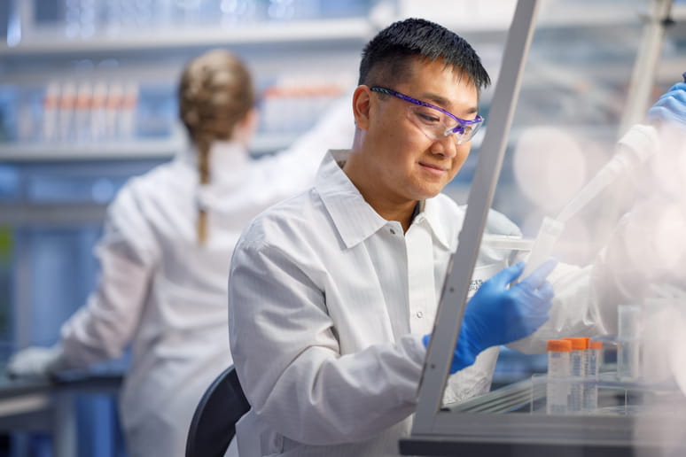 One lab technician filling a test tube and another working in the background.