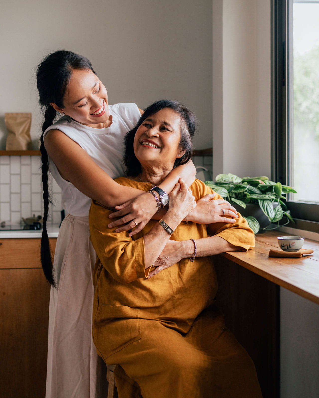 A woman hugging her mother in front of a sunny window.