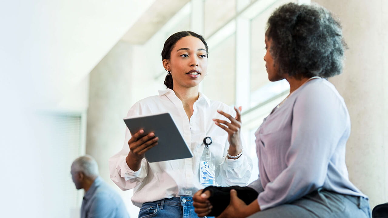A woman holding a tablet and speaking with a patient.