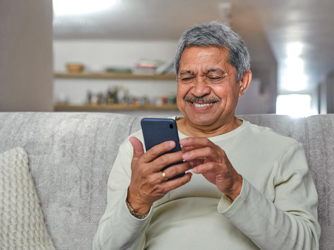 A man sitting on a couch holding a mobile device, looking at it smiling.