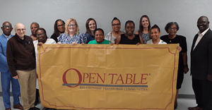 A diverse group of Exact Sciences and Open Table representatives smile while holding a large yellow banner reading “Open Table: Relationship Transforms Communities” against a gray wall.